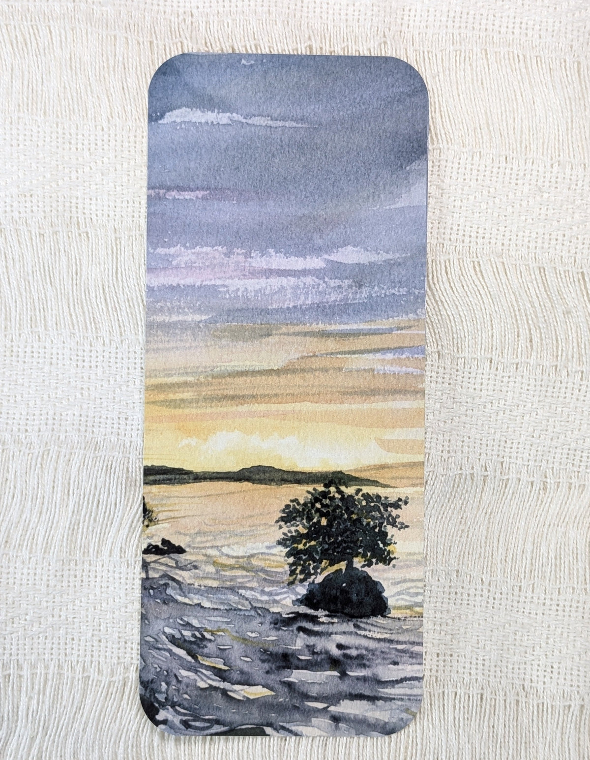 A rectangular bookmark with rounded corners of a watercolor painting of a coastal landscape with a lone mangrove tree on rocks at sunset.