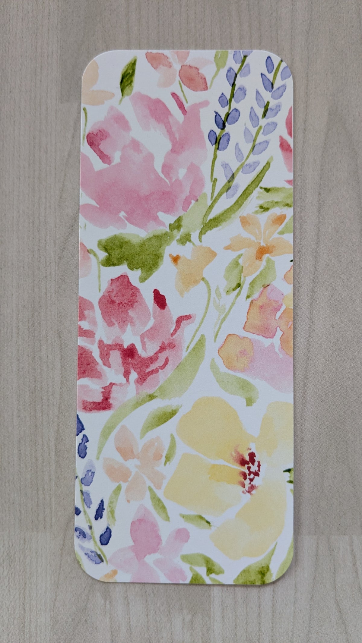 A rectangular bookmark with rounded corners featuring a colorful watercolor floral pattern, displayed flat on a light gray wood-textured surface.