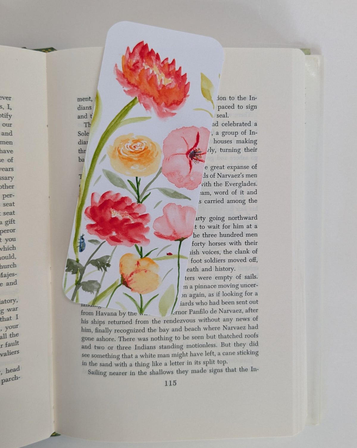A decorative bookmark featuring watercolor illustrations of red, pink, and orange flowers with green leaves, shown resting on the pages of an open book.