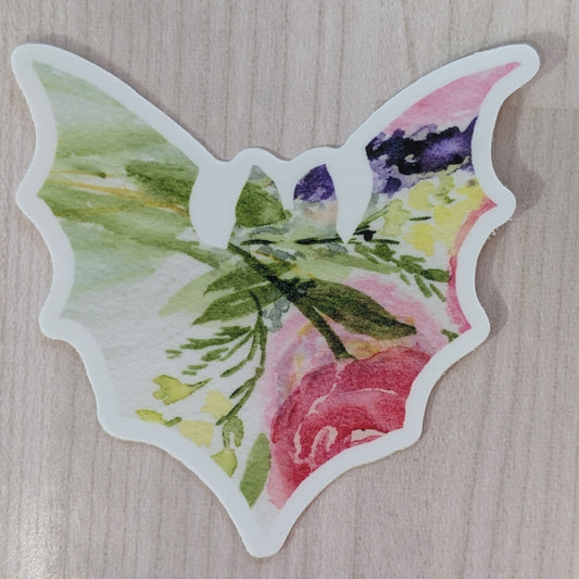 A bat shaped die-cut sticker featuring a watercolor floral design with pink, green, yellow, and purple tones, shown on a wood textured background.