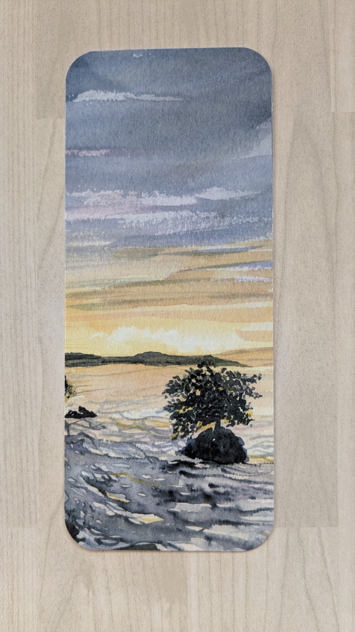 A rectangular bookmark with rounded corners of a watercolor painting of a coastal landscape with a lone mangrove tree on rocks at sunset.